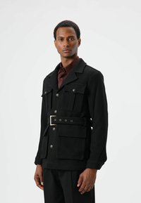 Zwarte gestructureerde jas met vier voorzakken, brede kraag en een riem met metalen gesp. Gecombineerd met een donker shirt.