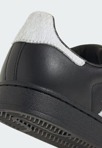 Čierna kožená teniska s textúrovanou gumovou podrážkou a mäkkým sivým umeleckým kožúškom na päte. Čistý dizajn s prešívanými detailmi.