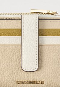 Κοντινή λήψη μιας μπεζ δερμάτινης τσάντας με χρυσό φερμουάρ, άσπρη ετικέτα, κίτρινη ρίγα και χρυσό λογότυπο Coccinelle.