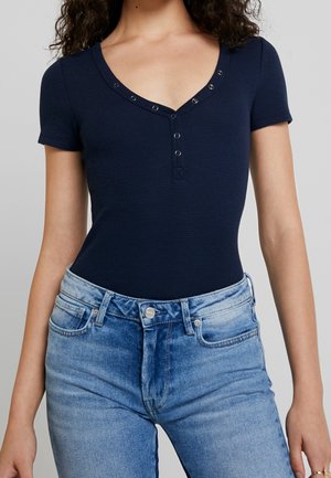 Marineblauw kortärmelig bodysuit gemaakt van geribd stof met een V-hals en drukknopsluiting. Draagt lichtblauwe hoge taille jeans.