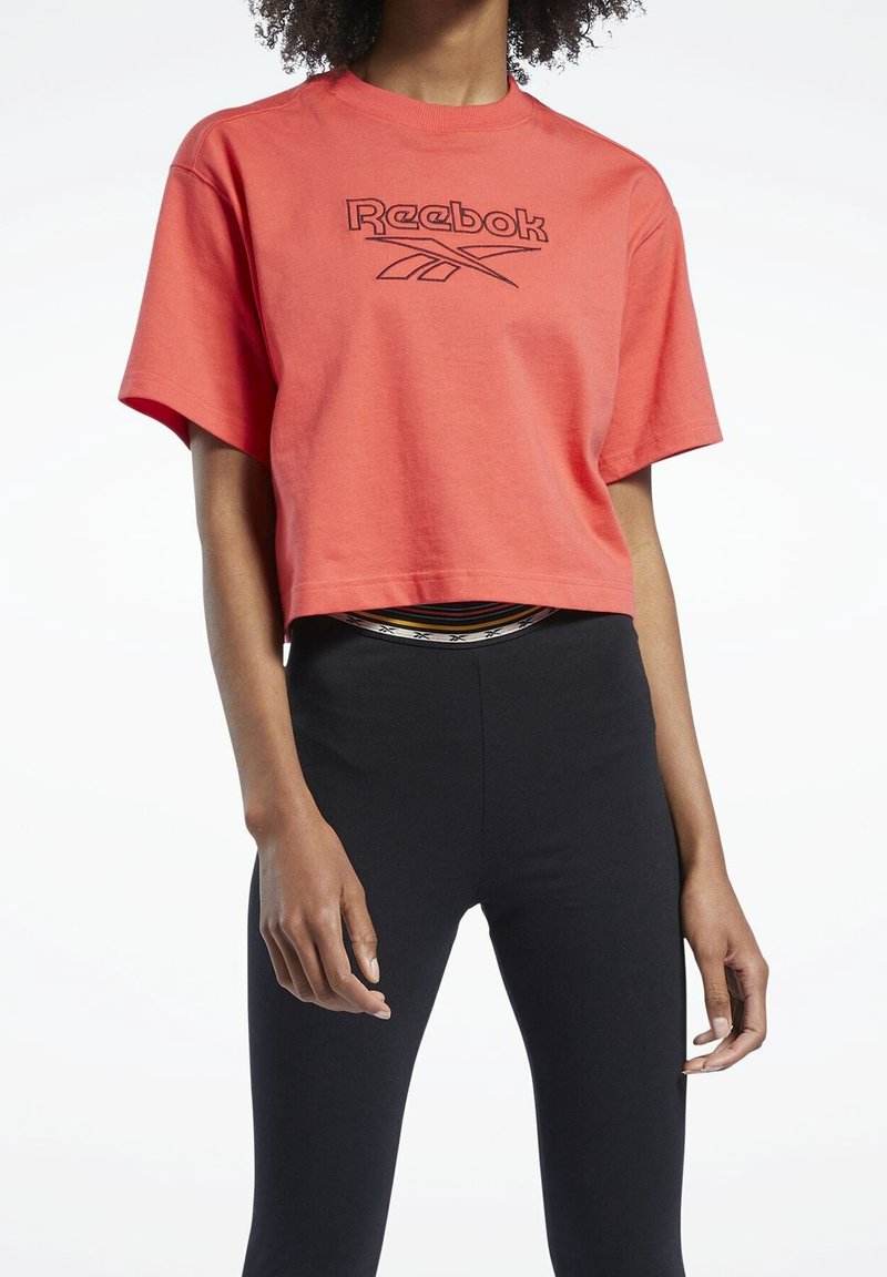Červené zkrácené tričko s černým logem Reebok na přední straně, v kombinaci s černými legínami. Měkká bavlněná tkanina a volný střih.
