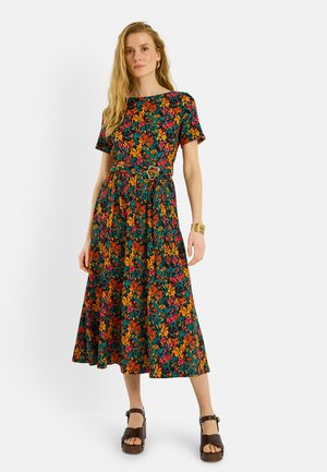 Mujer con un vestido midi de manga corta y cinturón, con un patrón floral colorido y sandalias plataforma marrones, posando frente a un fondo blanco.