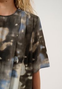 Průhledná blůza s tečkovaným vzorem v černé, modré a béžové barvě. Volný střih s krátkými, splývavými rukávy a vysokým výstřihem.