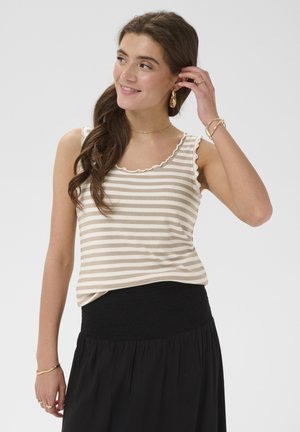 Femme aux longs cheveux bruns, portant un haut sans manches à rayures beige et blanc et une jupe noire, souriante et touchant sa boucle d'oreille.