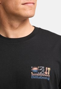 Musta puuvillapaita, jossa on värikäs grafiikka vasemmassa rinnassa yhdistettynä palmupuun ja geometrisiin kuvioihin, ja joka on varustettu "Billabong"-logolla.