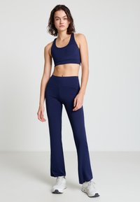 Frau trägt einen marineblauen Sport-BH und passende ausgestellte Leggings mit weißen Turnschuhen, steht vor einem einfarbigen hellen Hintergrund.