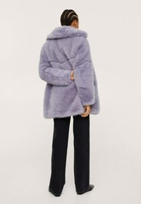 Persona con capelli intrecciati che indossa un cappotto in pelliccia sintetica lavanda, pantaloni neri e scarpe nere, in piedi con un braccio dietro la schiena.