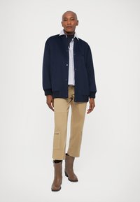 Námořnická vlněná bunda s knoflíkovým zapínáním, přeložená přes světle modrou pruhovanou košili a khaki cargo kalhoty, doplněná o hnědé kotníkové boty.