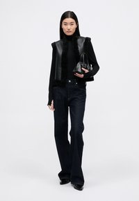 Μαύρο δερμάτινο κοντό γιλέκο με τριχωτή επένδυση, συνδυασμένο με μαύρο εφαρμοστό μανίκι που κλείνει στον λαιμό και σκούρους εκρού τζιν με φαρδιά γραμμή. Το μοντέλο κρατά μια μικρή μαύρη τσάντα.