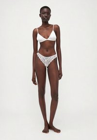 Bela satenska bralette z ozkimi naramnicami, v kombinaciji z belim čipkastim tangicami z nazobčanimi robovi, prikazana na modelu v stoječem položaju.