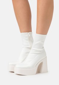 White heeled ankle boots with a smooth, shiny finish, featuring a zip closure and a chunky platform sole with a thick block heel.