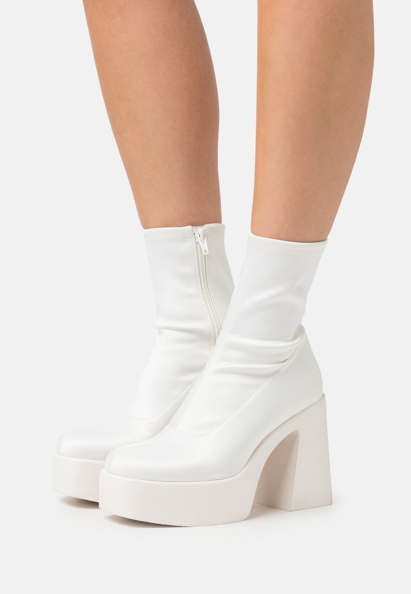 White heeled ankle boots with a smooth, shiny finish, featuring a zip closure and a chunky platform sole with a thick block heel.