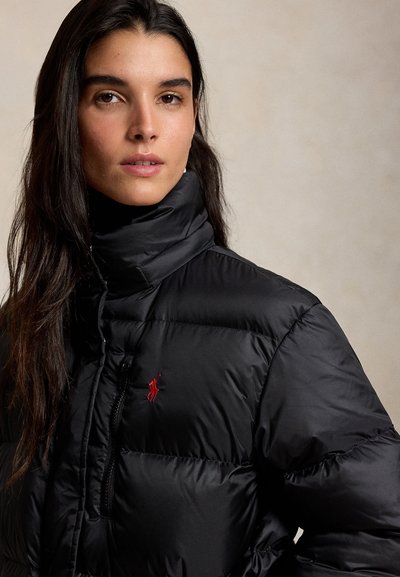 Mujer joven con cabello largo y oscuro que lleva una chaqueta acolchada negra con un logo rojo bordado en el pecho, sobre un fondo neutro.
