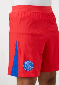 Røde atletiske shorts med vertikal ribbet tekstur, blå sidelinje og rundt blåt logo med "PARIS SAINT-GERMAIN" nederst.