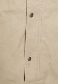 Tessuto beige con una trama testurizzata presenta bottoni a pressione metallici lungo la chiusura, enfatizzando un design durevole e funzionale.