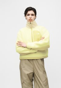 Άτομο με κοντά σκουρόχρωμα μαλλιά φοράει ανοιχτό κίτρινο φλις μπουφάν με φερμουάρ και μπεζ παντελόνι cargo, στέκεται με σταυρωμένα τα χέρια πάνω σε λευκό φόντο.