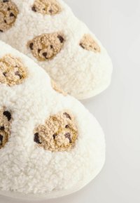 White fluffy slippers featuring bear faces in brown and tan, with a textured fabric surface and round shape. Soft and cozy design.