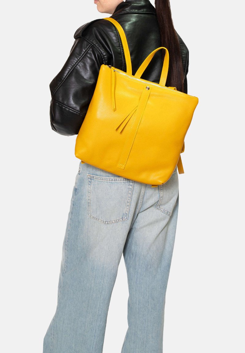 Zaino in pelle gialla con texture liscia, dotato di spallacci regolabili e chiusura con zip, indossato sopra una giacca nera e jeans azzurri chiari.