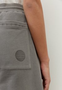 Jupe en coton grise avec une poche arrière, ornée d'un logo en cercle. Texture lisse et taille élastique.