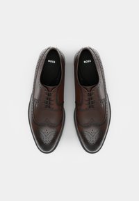Zapatos de brogue de cuero marrón con intrincados detalles de punta alada, perforaciones y un acabado suave, con una plantilla negra que lleva el logo 'BOSS'.