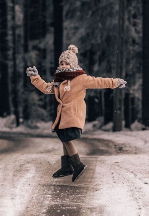 Cappotto in shearling marrone su una gonna scura, sciarpa rossa, guanti grigi e berretto lavorato a maglia con pom-pom. Stivali neri su una strada innevata in una zona boschiva.