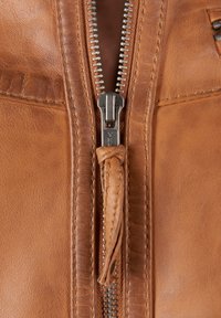 Close-up van een brown leren jack met een metalen YKK-rits en een gevlochten leren trekker, een gestructureerd oppervlak en zichtbare stiksels.