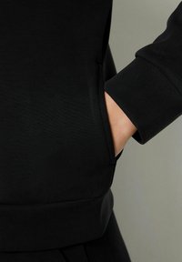 La mano di una persona è parzialmente all'interno della tasca laterale di un indumento nero a maniche lunghe con polsini e orlo a coste.