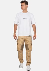 T-shirt blanc à manches courtes avec un logo noir associé à un pantalon cargo beige doté de poches zippées et d'accents côtelés sur les jambes.