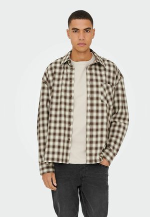 Bruin en crèmekleurig geruite flanellen shirt met lange mouwen, knoopsluiting aan de voorkant en een borstzak, gedragen over een lichtbeige t-shirt en donkere spijkerbroek.