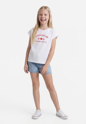 Šypsosi mergina su ilgais šviesiais plaukais, vilkinti baltą marškinėlį su grafiniu atspaudu, džinsinius šortus ir baltus sportbačius, stovinti ant balto fono.
