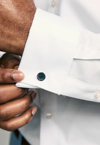 Bijela košulja sa teksturiranim završetkom na manžeti, koja sadrži crnu heksagonalnu manžetnu. Ton kože je vidljiv dok se drži manžeta.