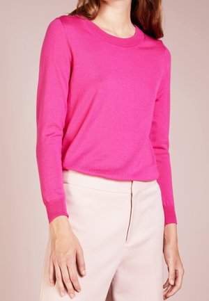 Kvinde iført en knaldrød langærmet sweater og lyse beige bukser mod en ensfarvet baggrund, med armene afslappet ned langs siderne.