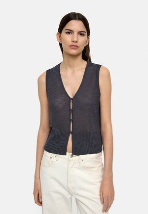 Młoda kobieta o ciemnych, prostych włosach, ubrana w bezrękawnik dzianinowy w kolorze węgla z guzikami oraz kremowe spodnie.