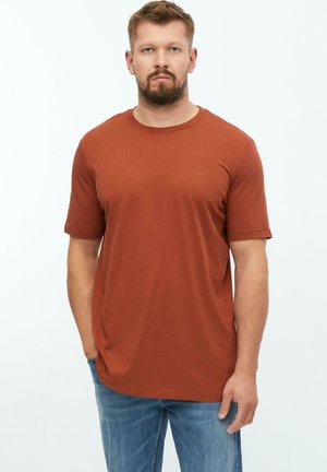 Mann mit Bart, der ein rostfarbenes T-Shirt mit kurzen Ärmeln und blaue Jeans trägt, steht vor einem schlichten weißen Hintergrund.