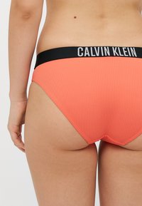 Kalvinas Klein ribotu apatinio trikotažo rinkinys koralinės spalvos, su juodu logotipu pažymėtu juosmeniu. Galinis vaizdas rodo elastingą medžiagą ir prigludusį siluetą.
