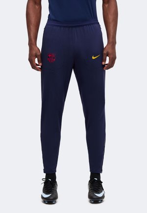 Homme portant un pantalon de sport bleu marine avec un logo rouge du FC Barcelone et un swoosh jaune Nike, assorti à des crampons de football noirs.