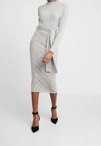 Femme portant une robe midi grise ajustée à manches longues avec une ceinture à la taille, associée à des escarpins noirs à bride de cheville, debout devant un fond blanc uni.