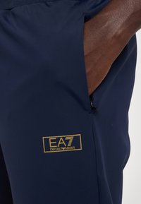 Tamsiai mėlynos sportinės šortai, pagaminti iš lygaus audinio, su užtrauktuku užsegamu kišeniu. Priekyje yra auksinė "EA7 Emporio Armani" logotipo etiketė.