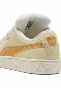 Barna velúr sneaker narancs akcentussal, texturált talppal és párnázott nyelvvel. A sarokon logó található, valamint szőtt részlet az oldalon.