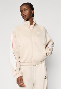 Μπεζ αθλητικό μπουφάν με φερμουάρ, με κόκκινες και λευκές λεπτομέρειες, ριμπ μανσέτες και απαλό ύφασμα. Διαθέτει λογότυπο Adidas στο στήθος.
