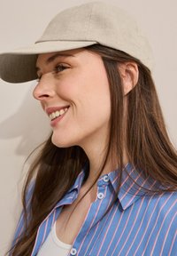 Gorra beige con visera curva, llevada con una camisa a rayas azul y rosa con botones, delante de un fondo neutro.