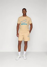 T-shirt Lotto bege com um design gráfico azul e laranja, combinada com calções a condizer. O tecido aparenta ser macio; o modelo usa ténis brancos.