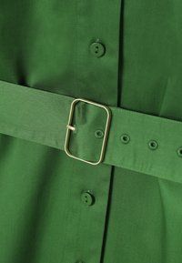 Detailný záber na zelenú látku so zodpovedajúcim opaskom s obdĺžnikovou zlatou prackou a zelenými gombíkmi, ktoré sú na odeve prišité vertikálne.