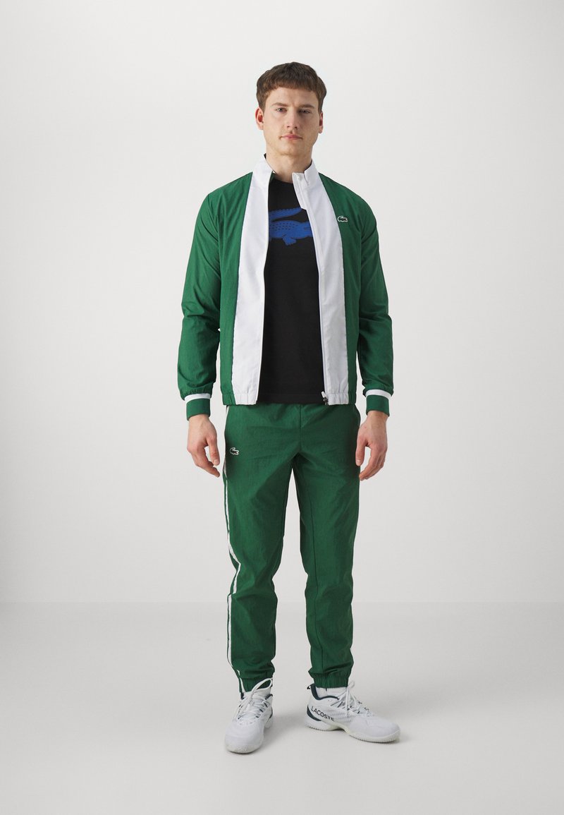 Zielona kurtka sportowa z białym zamkiem i wykończeniem, zestawiona z pasującymi zielonymi spodniami z białymi paskami po bokach. Czarna koszulka z niebieskim wzorem.