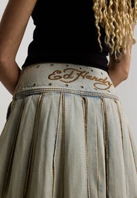 Close-up of person wearing light denim skirt with "Ed Hardy" embroidered on waistband and black sleeveless top, showing blonde curly hair.