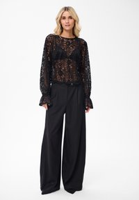 Kobieta w przezroczystej czarnej koronkowej bluzce z długimi rękawami i szerokich czarnych spodniach, stojąca na jednolitym białym tle.