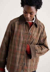 Brūns kreklu uzvalks ar pārklājumu un aprocēm no korduroja, pogu aizdari un brīvu piegriezumu. Zem tā ir redzams sarkans teksturēts džemperis.