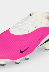 Άσπρο και ροζ ποδοσφαιρικό παπούτσι Nike με μαύρο λογότυπο Swoosh, λευκά κορδόνια και διάφανα πέλματα, εμφανίζεται από γωνία σε ανοιχτόφωτο φόντο.