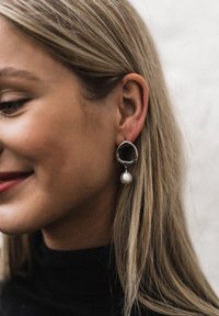 Orecchini a cerchio in argento con una forma organica, caratterizzati da una grande perla bianca che pende da ciascun orecchino. Indossati da una persona con capelli chiari.