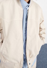 Blouson aviateur beige avec fermeture éclair à l'avant, poignets côtelés et poches latérales, porté sur une chemise à boutons bleu clair. Tissu texturé.